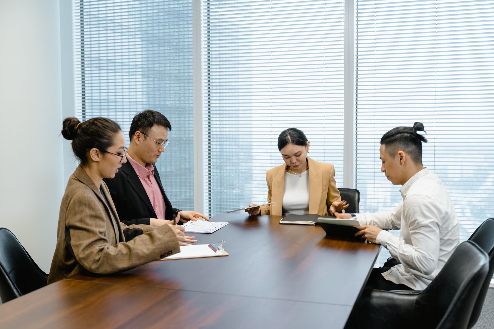 Five colleagues in discussion around a wooden meeting table in an office with window blinds.
